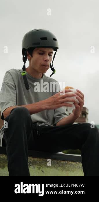 A teenage boy in a helmet eats a sandwich while taking a break at a ...
