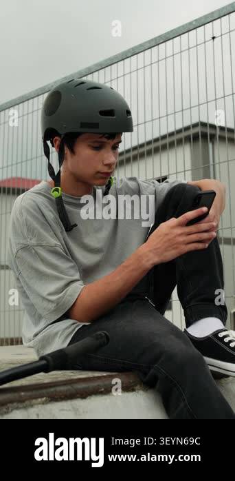 A teenage boy in a helmet sits on a ramp at a skatepark and uses his ...