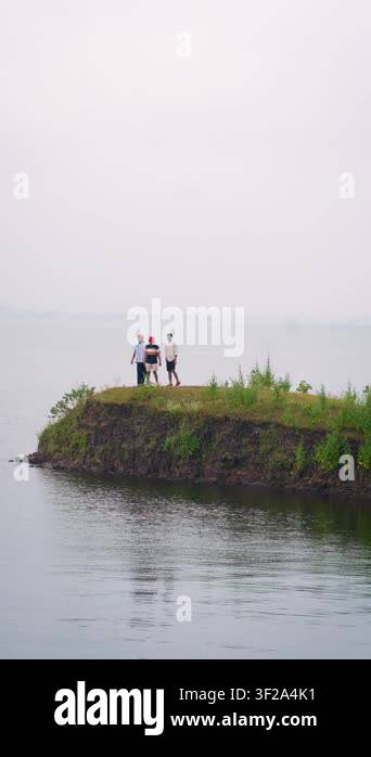 Three people stand on a small grassy island surrounded by calm water in ...