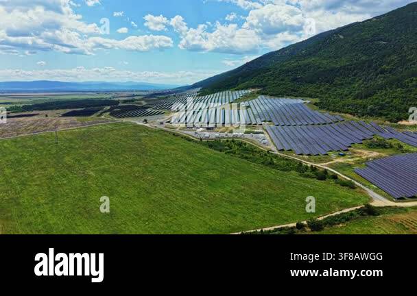 Large solar panels are arranged in rows on a hillside. Green fields are ...
