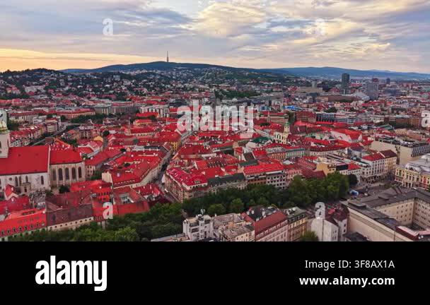 The city shows buildings with red roofs and green trees. The sun sets ...