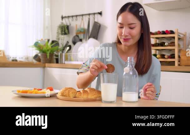 Young woman dipping a cookie into a glass of milk at a bright kitchen ...