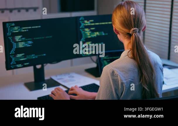 Woman programming on multiple monitors in a dimly lit workspace while ...