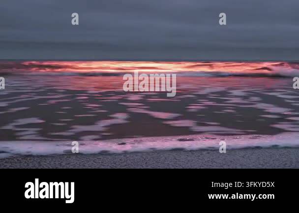 Gentle waves with pink sunset reflections roll onto a sandy beach under ...