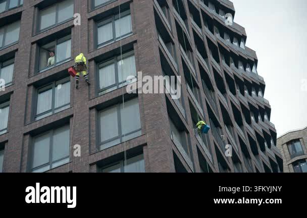 Two workers hang on ropes along tall brick facade, one near upper ...