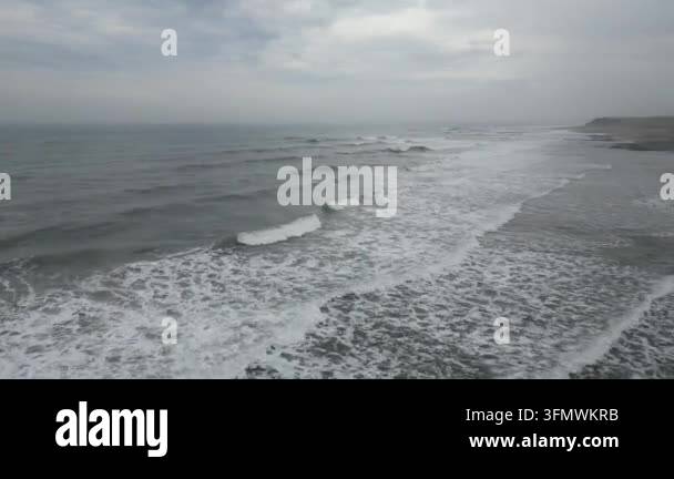 Aerial view of ocean waves crashing on a sandy beach under a cloudy sky ...
