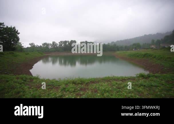 A wide shot of a calm lake surrounded by green grass and trees under a ...