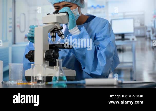 A scientist in a blue lab coat and mask is looking through a microscope ...