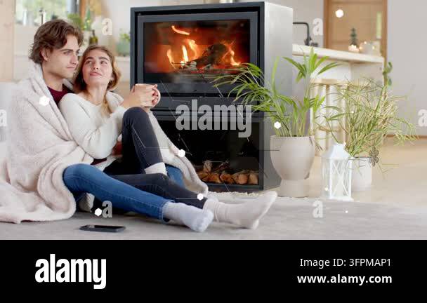 Couple cuddling under throw by stove phone buzzing, woman with mug ...