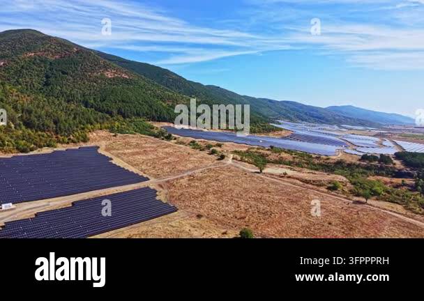 A solar energy farm covers a large area near hills and a water body ...