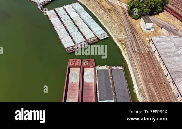 Several barges are lined up at a river dock. A railway track runs ...