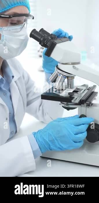 Female scientist analyzing sample through microscope, wearing ...