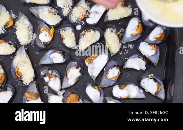 A hand uses a spoon to sprinkle grated cheese onto rows of mussels ...