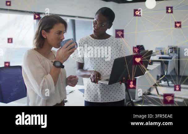 Two women working sipping lowering blue mug pointing at laptop ...
