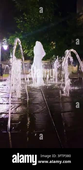 Illuminated city fountain water jets spray up in a square at night with ...