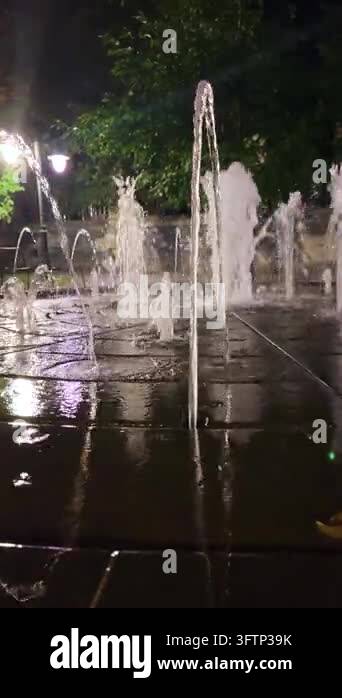 Illuminated city fountain water jets spray up in a square at night with ...