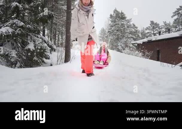 A woman pulling a child on a plastic sled down a snow-covered hill in a ...