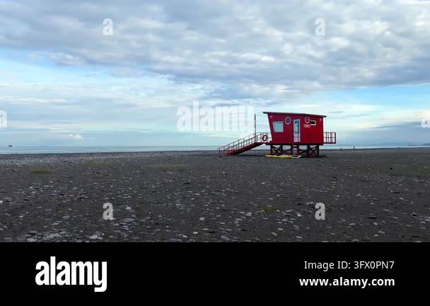 A red rescue hut stands beside the pebble shoreline, representing beach ...