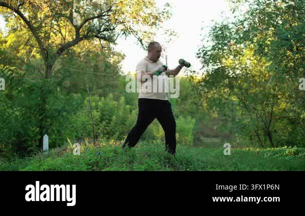 A man exercises in a sun-drenched forest, lifting dumbbells alternately ...