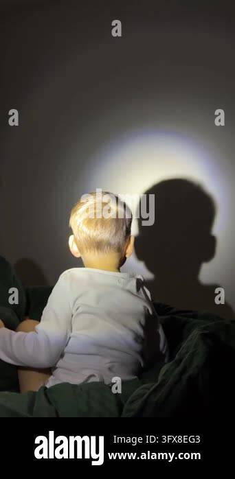 A toddler boy turns and moves on a bed while observing his silhouette ...