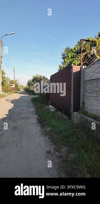 First-person POV walking along a narrow rural dirt road between garden ...