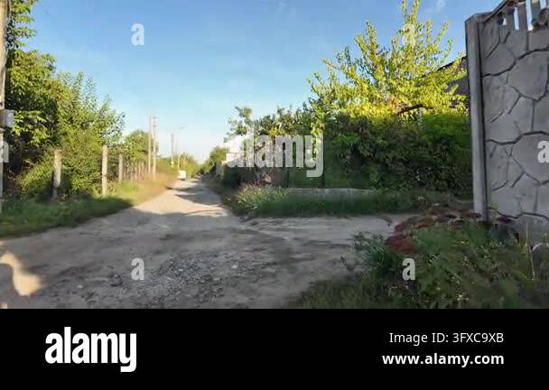 First-person POV walking along a narrow rural dirt road between garden ...