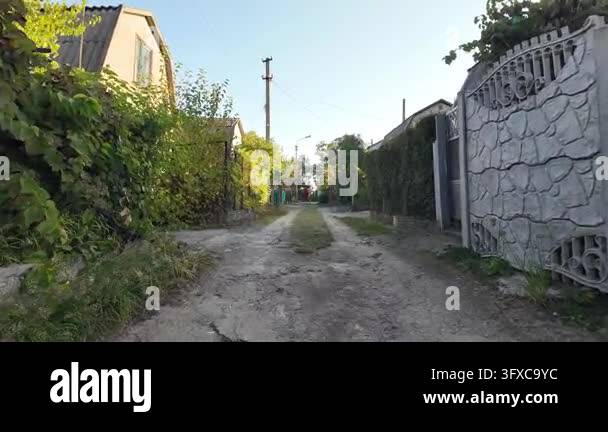 First-person POV walking along a narrow rural dirt road between garden ...