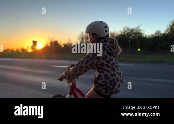 Sundet Line Model Child rides bicycle along quiet road at sunset, wearing white helmet ...
