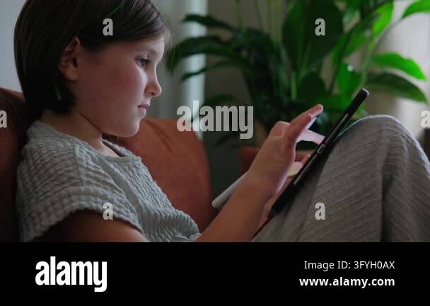 Side view of a focused young girl sitting on a sofa at home, using a ...