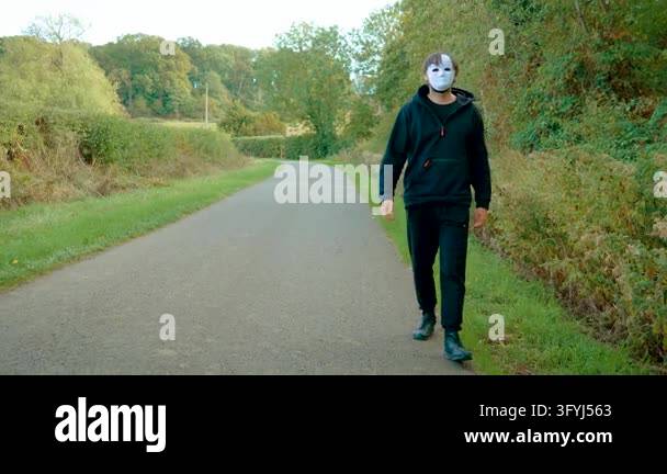 Criminal masked man walking along empty rural road in black hoodie and ...