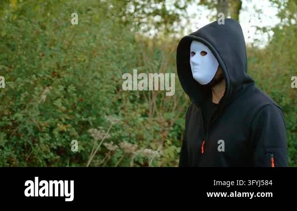 Criminal in white mask walking beside woodland in black hooded clothing ...