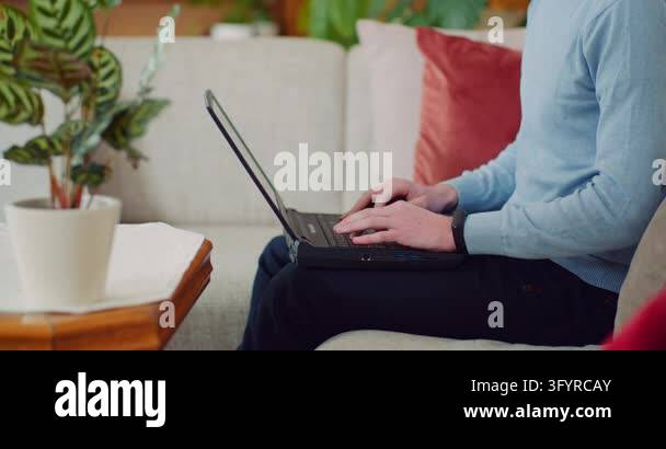 An adult sits on a couch and types on a laptop in a comfortable home ...
