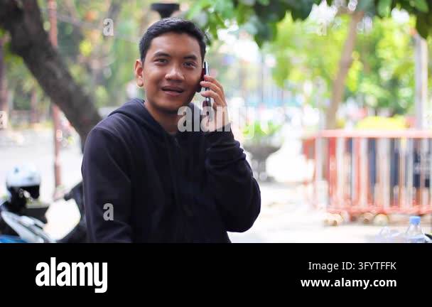 Asian man in a black hoodie holds a smartphone and talks on the phone ...