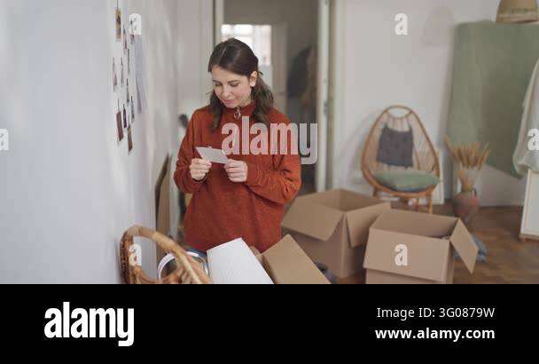 Brunette girl takes photos off wall and looks at them while moving in ...