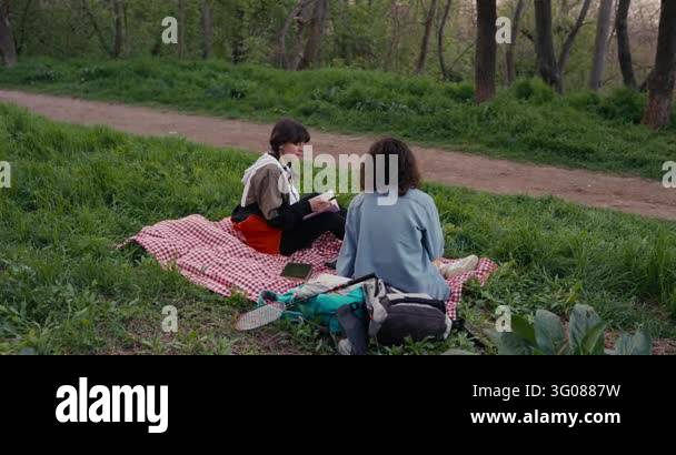 Two young women sit on red gingham blanket in leafy park clearing, one ...