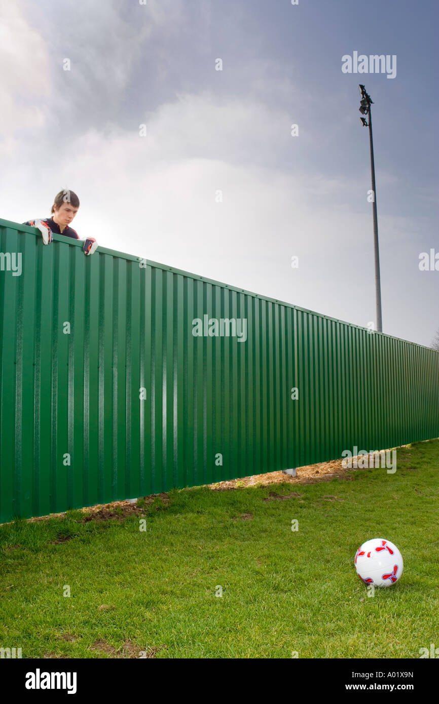 Young man wearing goalkeeper gloves looking over fence at soccer ball Stock Photo