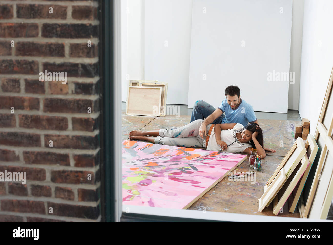 Couple reclining by painting on floor of studio, view through window
