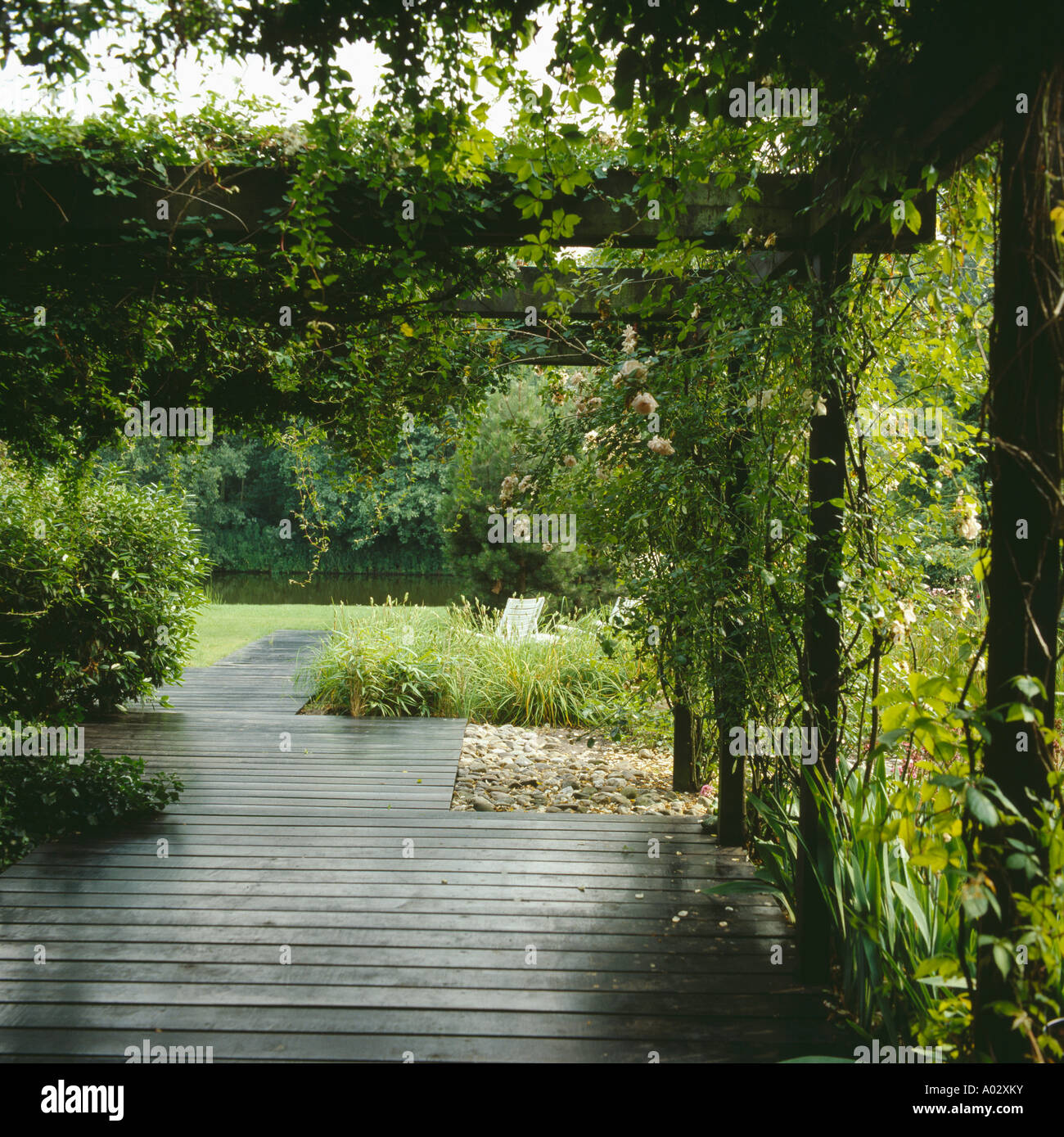 Decking path through a rose covered pergola to a lawn seating area and still water Stock Photo