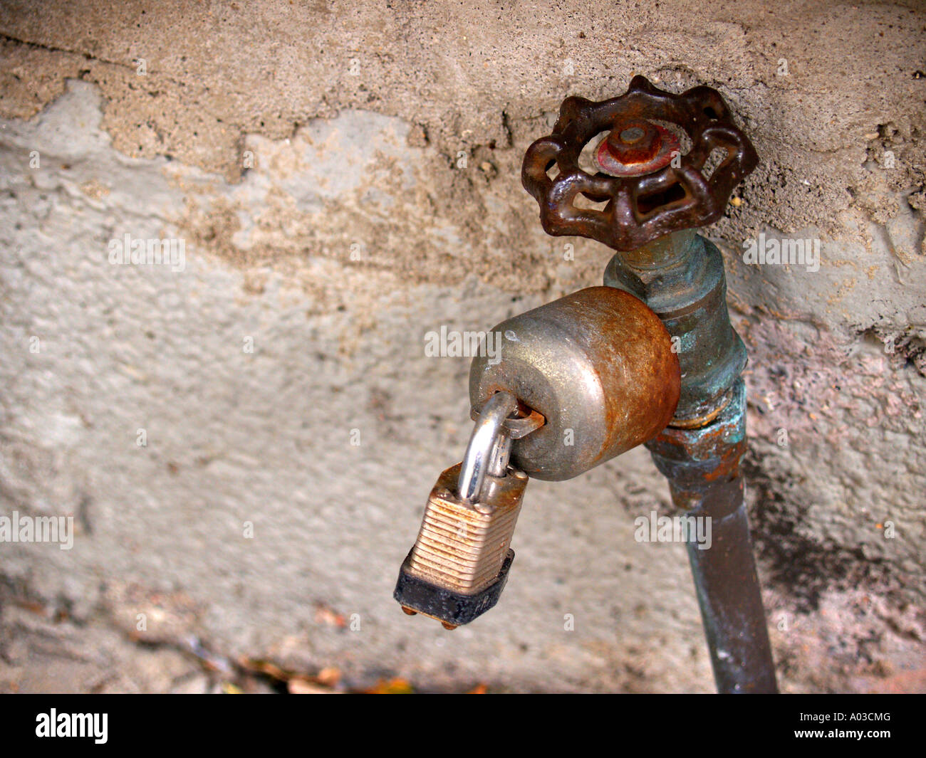 Rusted, padlocked water faucet in front of a speckled concrete wall. Stock Photo