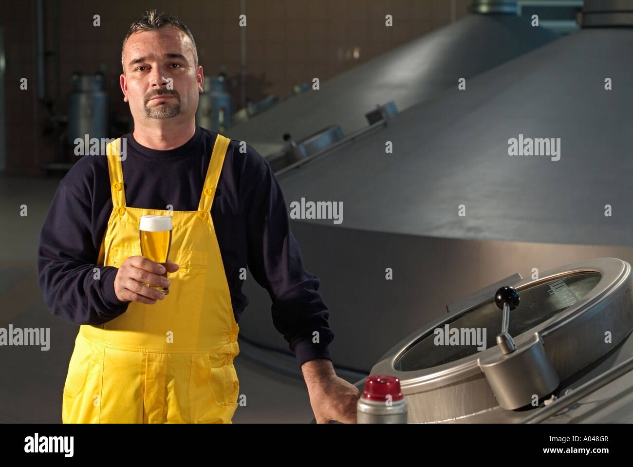 Brewery Worker Holding a Glass of Beer Whilst Standing Next to a Large Fermentation Vat of a Brewery Stock Photo
