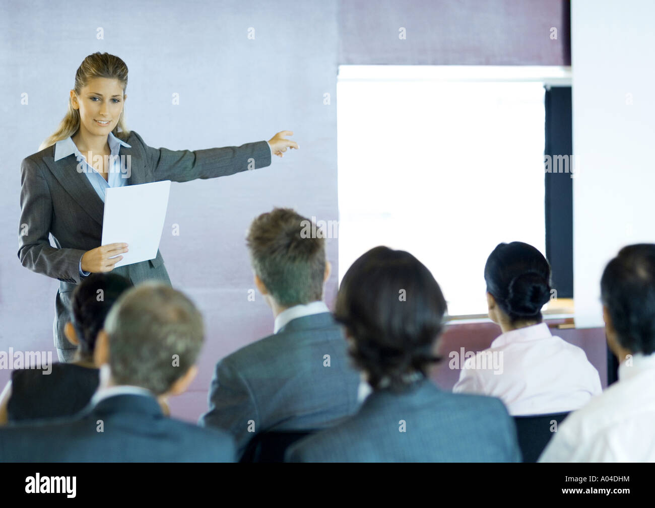 Executives sitting in seminar, woman standing facing group, gesturing to screen Stock Photo