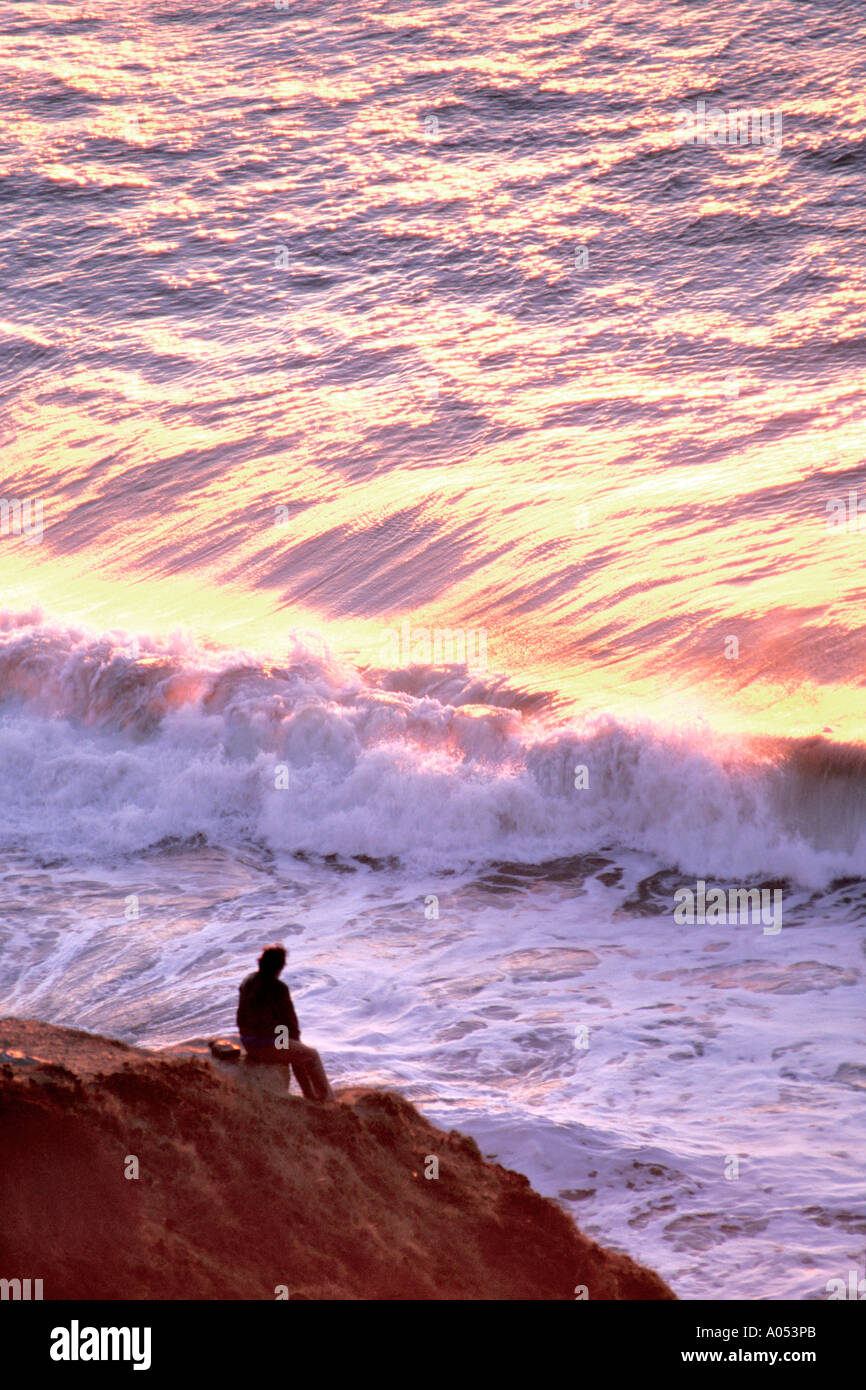 man sitting on rocky shorline watching the waves from the ocean crash on shore at sunset Stock Photo