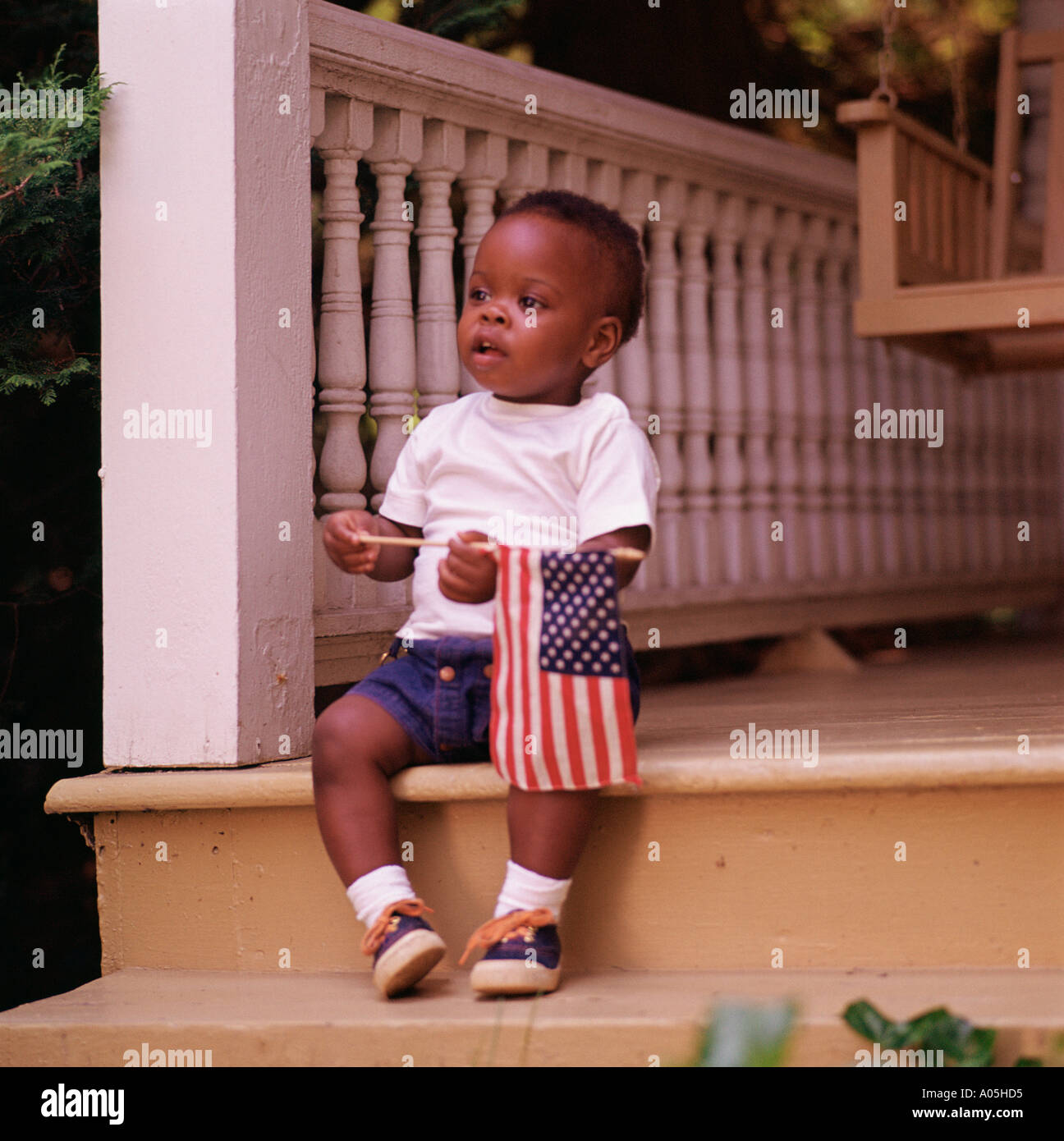 An African American toddler sitting on the steps of the front porch holding a small American flag Stock Photo