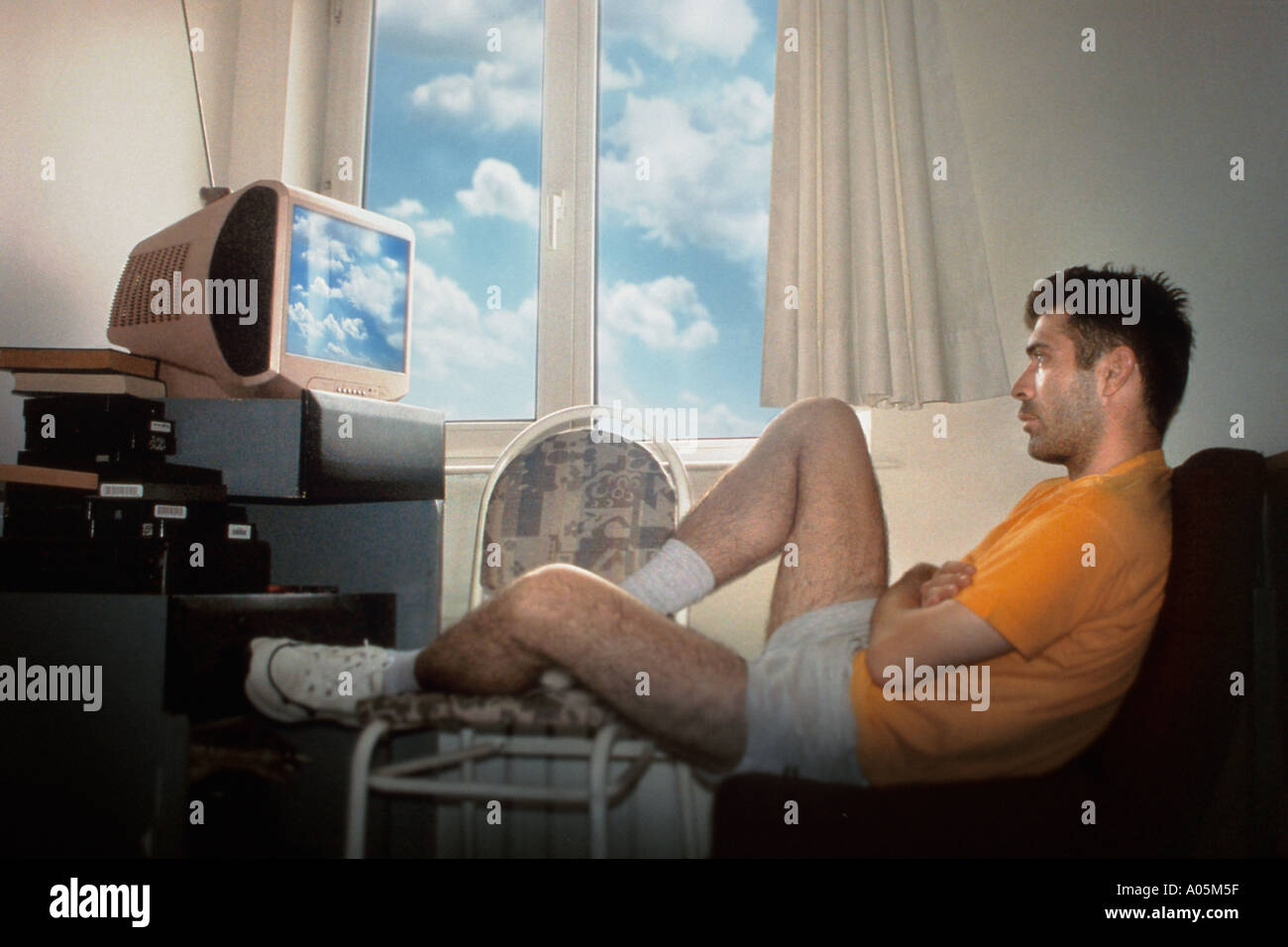 Man watching tv by himself with the window open and the clouds reflecting on the screen Stock Photo