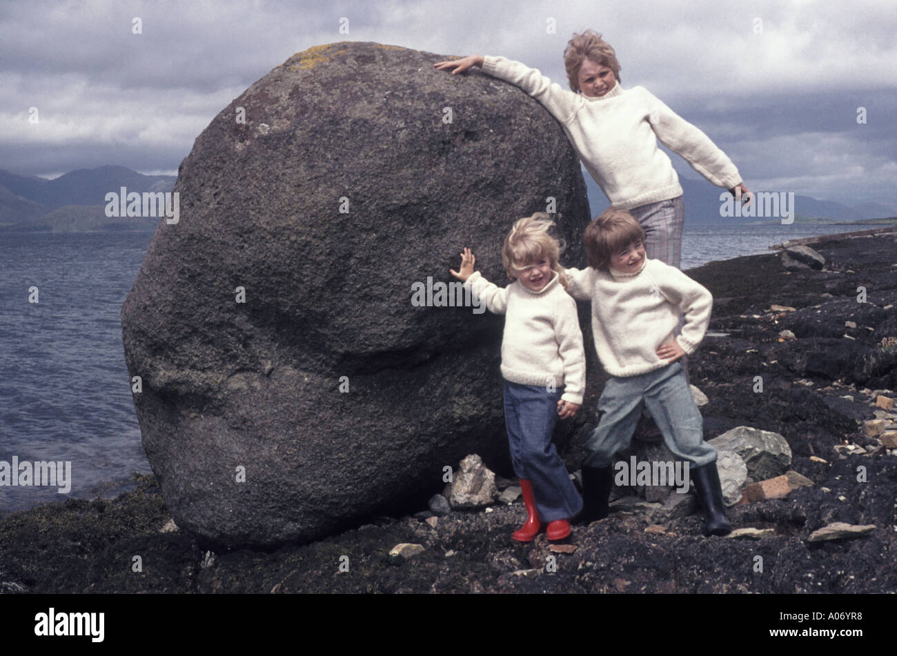 Scotland windy loch side pose 3 children in Aran jumpers on Scottish touring holiday in 1970’s pushing waterside boulder for the family photo album UK Stock Photo