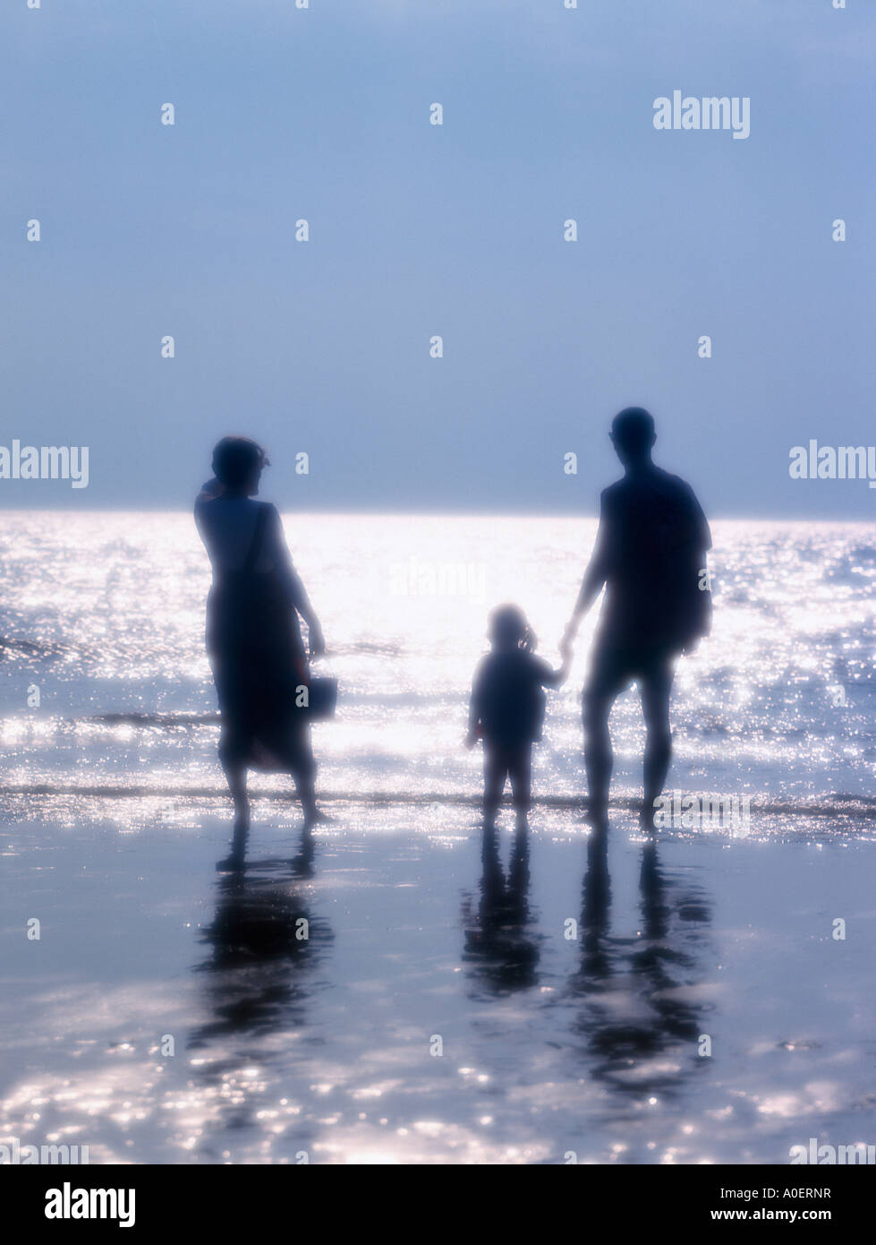 YOUNG COUPLE AND LITTLE GIRL ON BEACH AT WATERS EDGE IN SILHOUETTE AGAINST SPARKLING SEA AT MID DAY UK Stock Photo