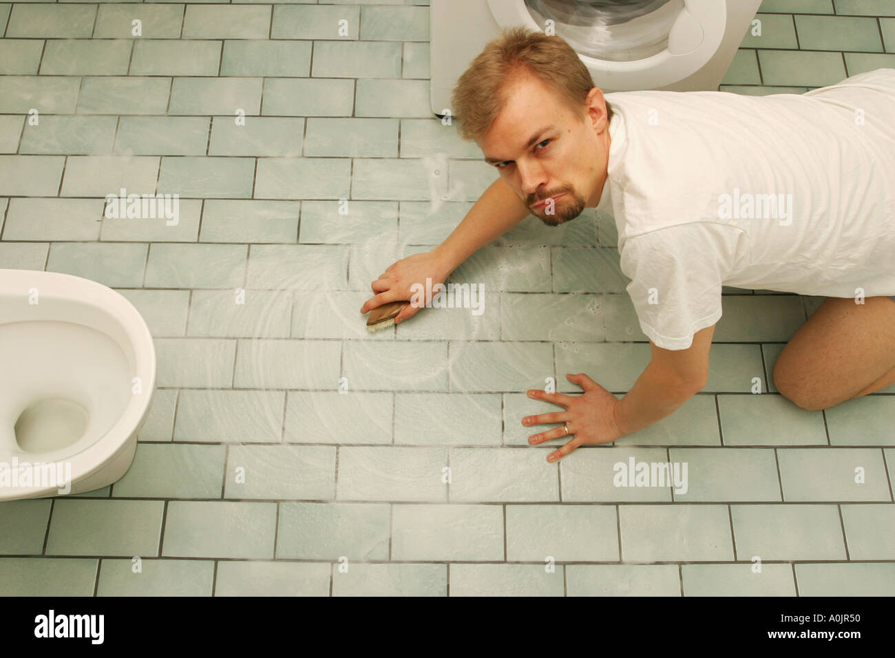 Grumpy man, 35 years old, scrubbing the bathroom floor, standing on his knees Stock Photo