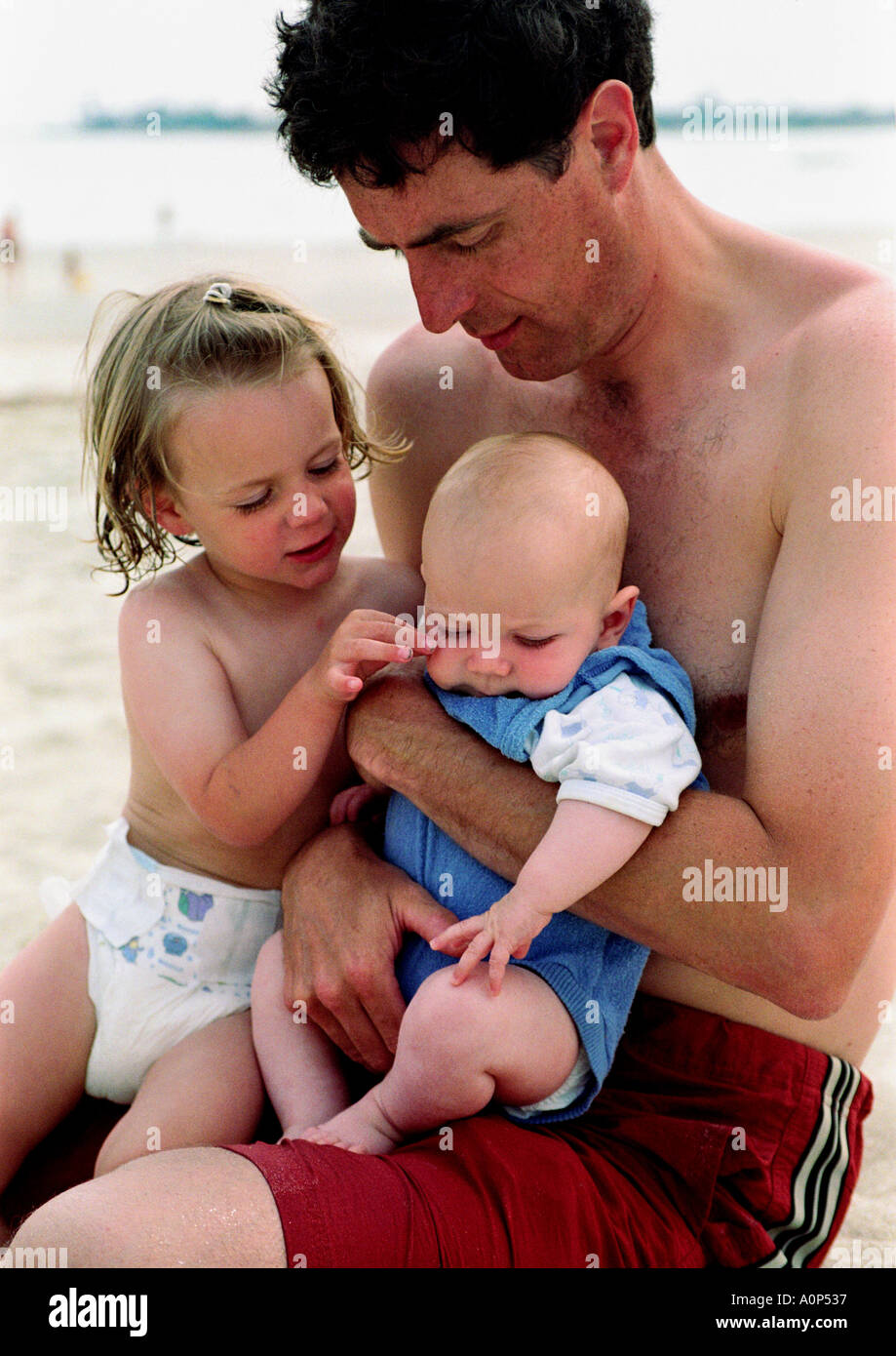 father holding new born baby at the beach on holiday, with sister pinching the baby's cheek Stock Photo