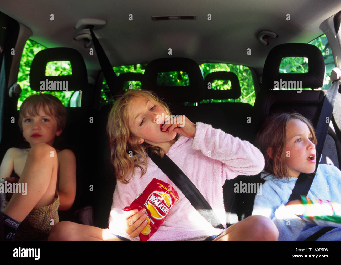 three young children in the car with seatbelts on and eating crisps Stock Photo