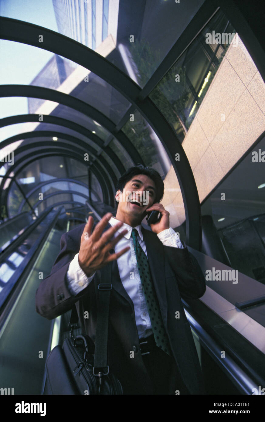 Asian American businessman talking on his mobile phone and gesturing as he rides down and outdoor escalator Stock Photo